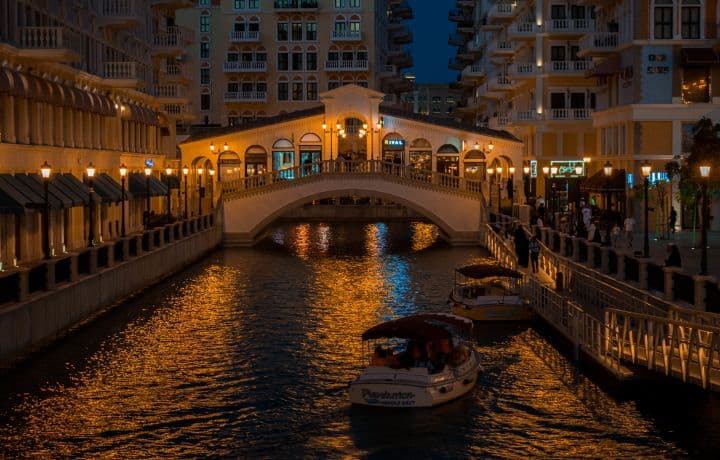 This one really gives off Venice vibes with the bridge, canal, and boats. The warm glow from the lights reflecting on the water makes it feel lively, especially with people walking around the area. Looks like a great spot to just take in the atmosphere.