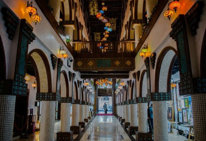 This image captures the interior of an art museum in Souq Waqif, Qatar, showcasing stunning traditional Arabian architecture. The arched corridors, intricate wooden details, and vibrant hanging lanterns create a warm and inviting ambiance, reflecting Qatar's rich cultural heritage.